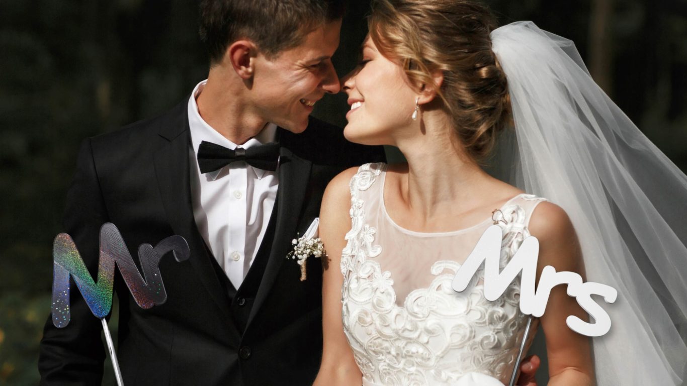 Brautpaar im Freien, lächelnd, mit "Mr" und "Mrs" in bunten Buchstaben im Vordergrund.