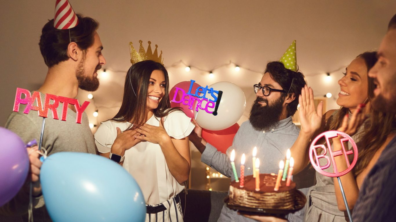 Gruppe von Menschen feiert mit einem Kuchen, Ballons und Partyhüten.